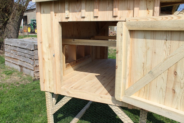 The side of the stable of a chicken tractor with the door standing open, with 3 laying nests and 2 perches inside.